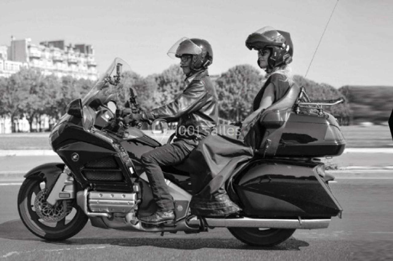 Deux personnes en moto de tourisme noire circulant sur une route bordée d'arbres et de bâtiments en arrière-plan.