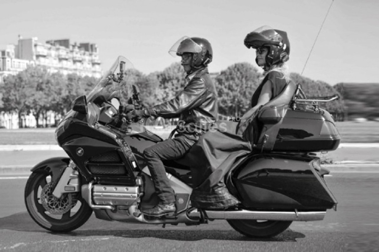Deux personnes en moto sur une route, portant des casques et des vêtements de protection, avec des arbres en arrière-plan.