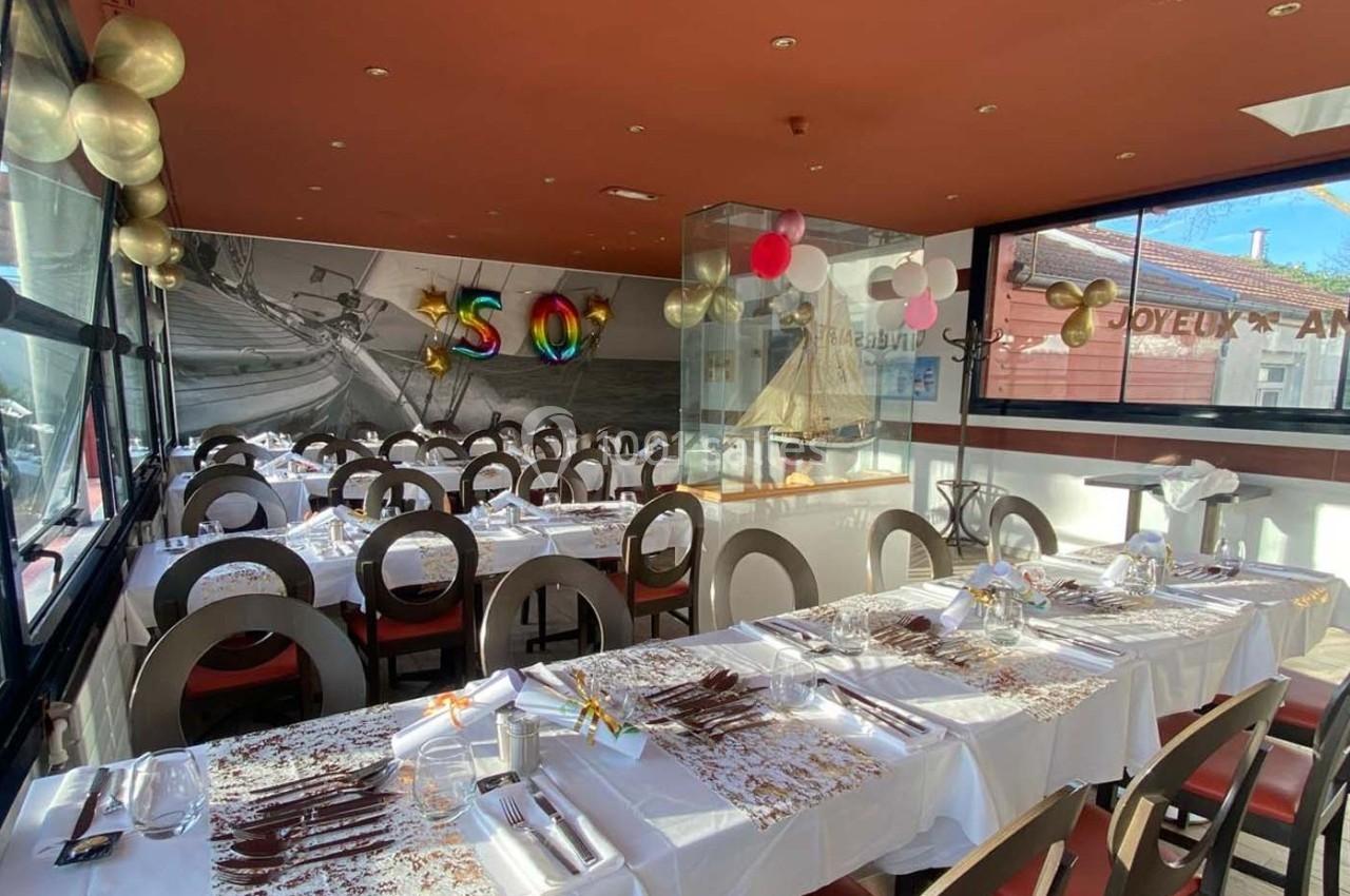 Salle de restaurant décorée pour un anniversaire avec ballons, nappes blanches et vue sur l'extérieur ensoleillé.