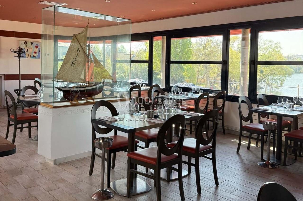 Salle de restaurant lumineuse avec des tables dressées, une maquette de voilier sous verre et vue sur un paysage extérieur.