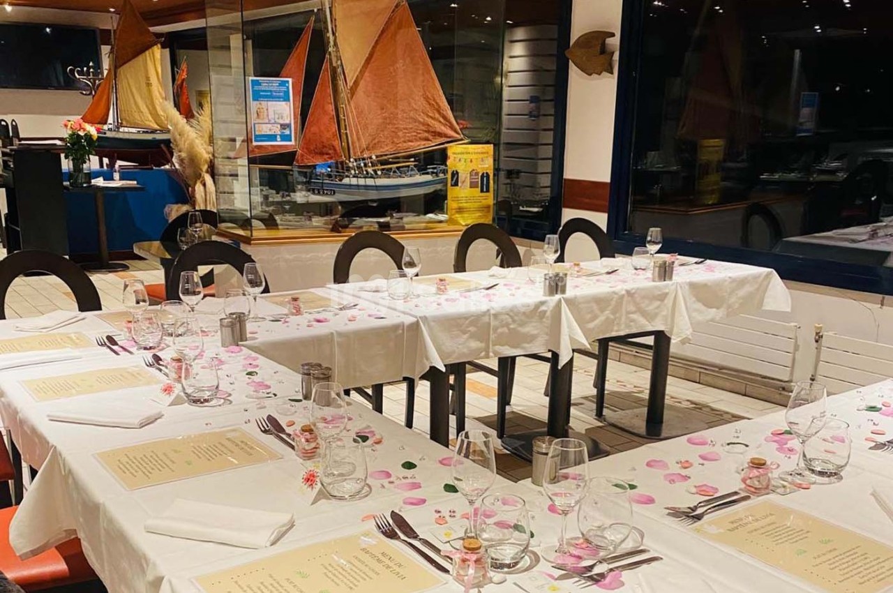 Salle de restaurant avec tables dressées, nappes blanches, menus et décoration maritime en arrière-plan.