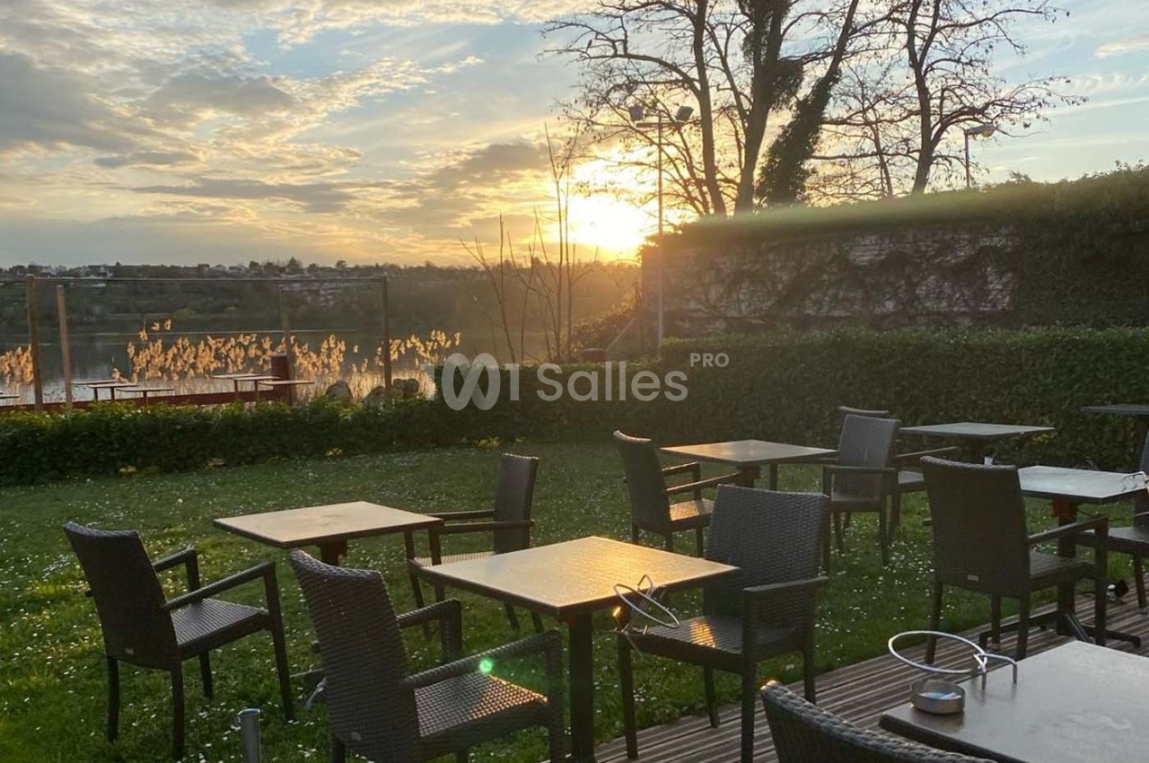 Terrasse extérieure avec des tables et des chaises en rotin, vue sur un lac au coucher du soleil.