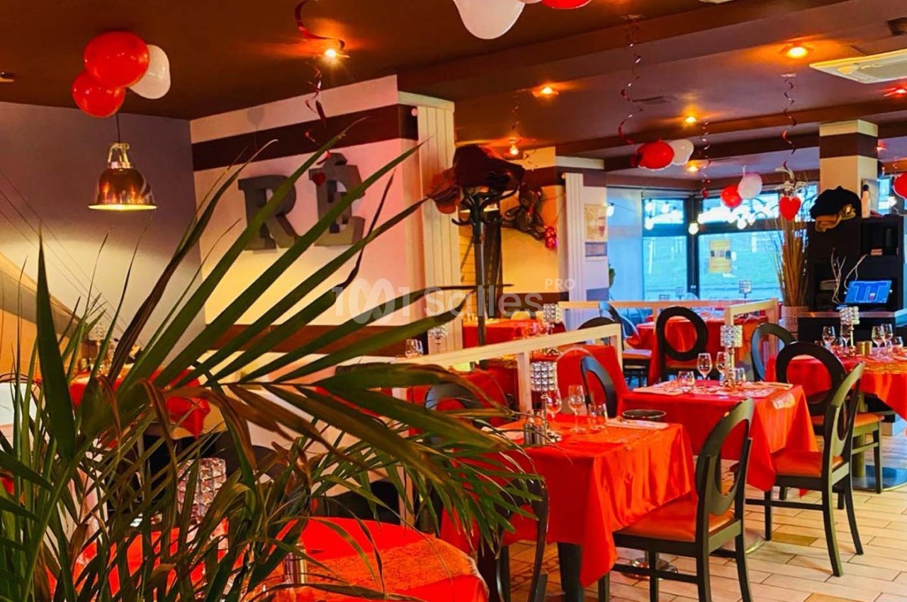 Salle de restaurant décorée avec des nappes rouges, des ballons suspendus et une lumière chaleureuse.