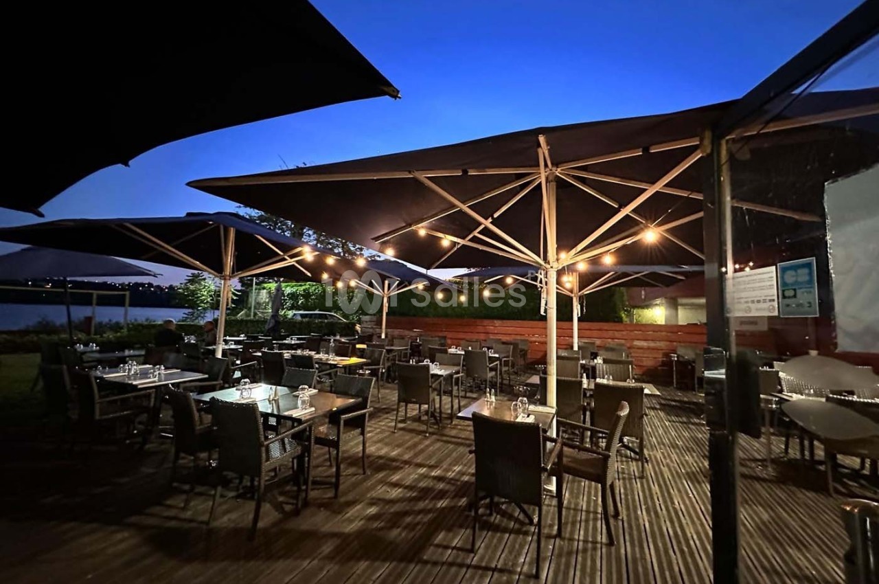 Terrasse en bois avec tables et chaises sous des parasols, éclairée par des guirlandes lumineuses au crépuscule.