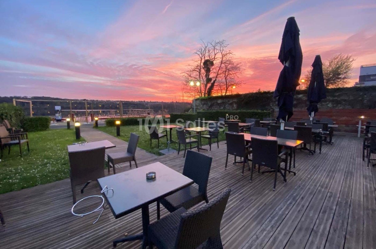 Terrasse en bois avec tables et chaises vides, vue sur un coucher de soleil coloré et un paysage dégagé.