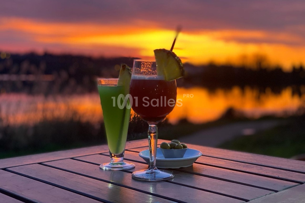 Deux cocktails colorés et une assiette d'olives sur une table en bois, avec un coucher de soleil en arrière-plan.