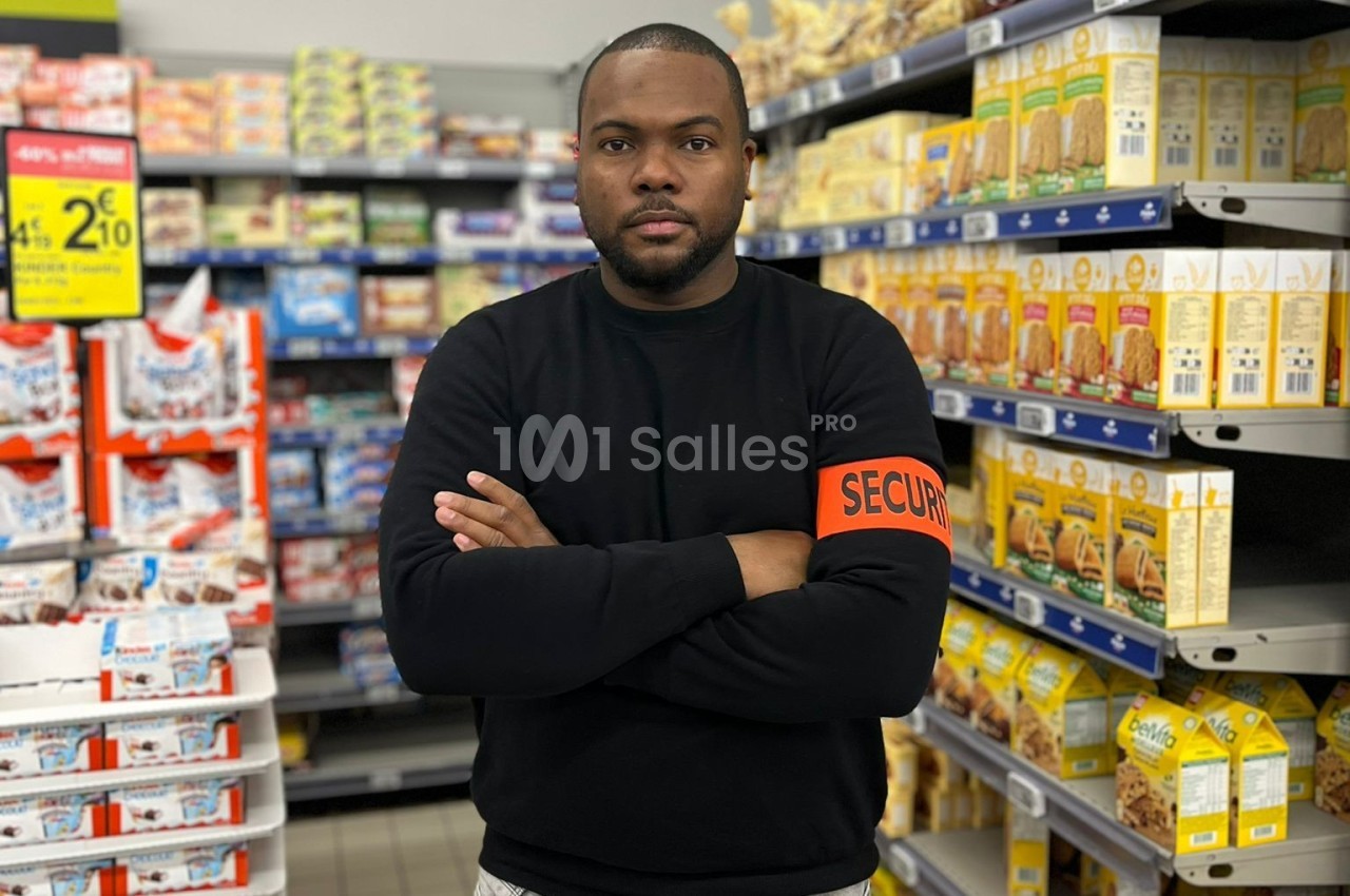 Un agent de sécurité en uniforme noir avec brassard orange se tient dans une allée d'un supermarché.