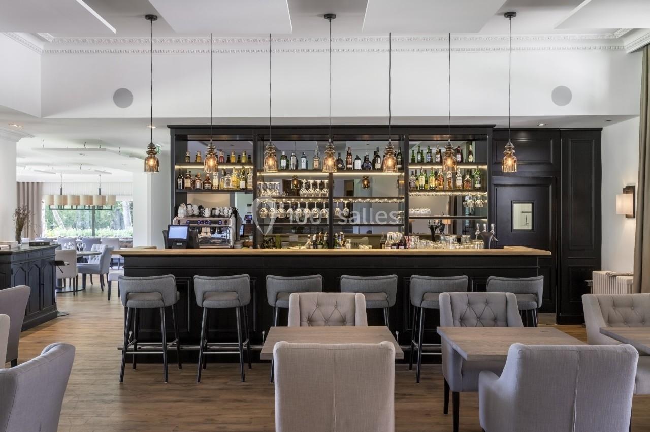 Bar moderne avec comptoir en bois, étagères garnies de bouteilles et verres, chaises hautes et tables en avant-plan.