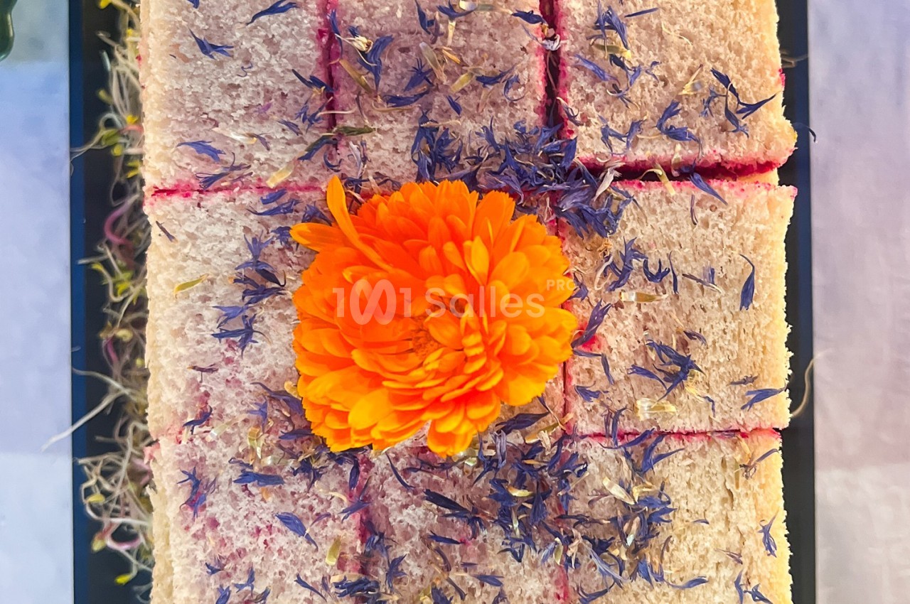 Gâteau rectangulaire décoré de fleurs séchées violettes et d'une fleur orange centrale, présenté sur un plateau noir.