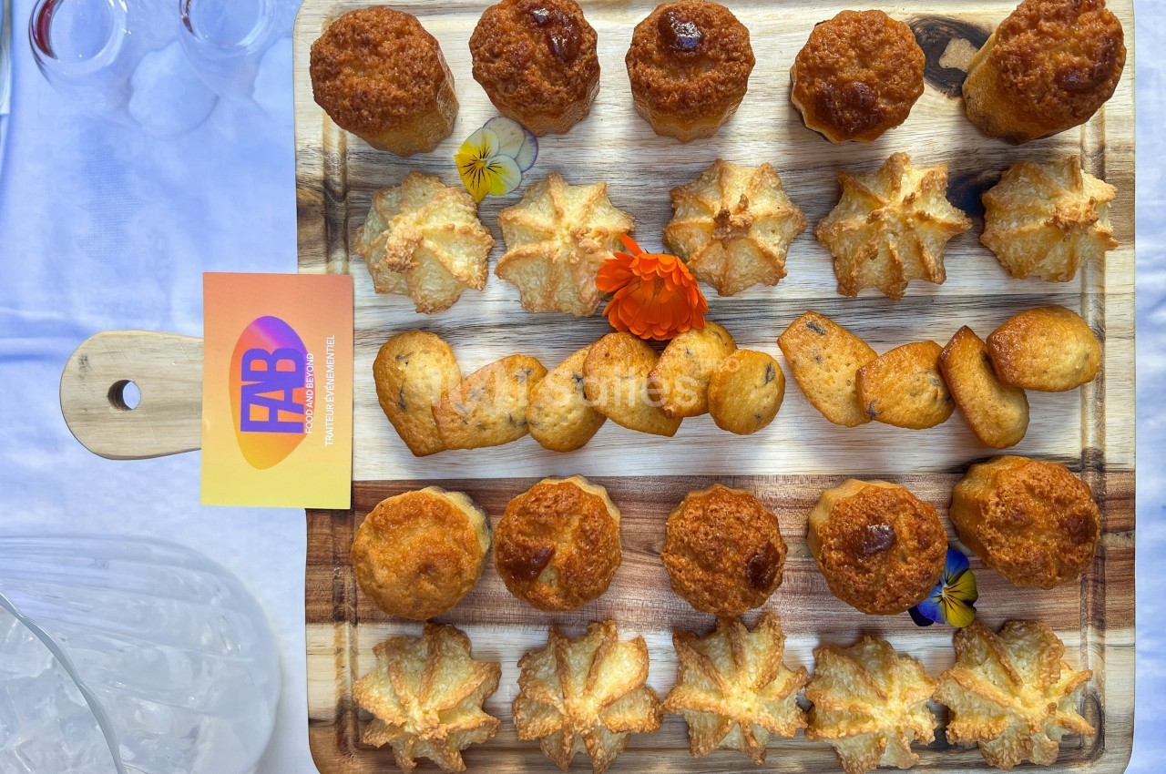 Plateau en bois garni de petits fours variés, dont des cannelés et des biscuits, décoré de fleurs comestibles.