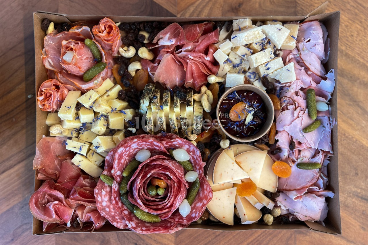 Plateau de charcuterie et fromages variés avec cornichons, fruits secs, noix et confiture dans une boîte en carton.