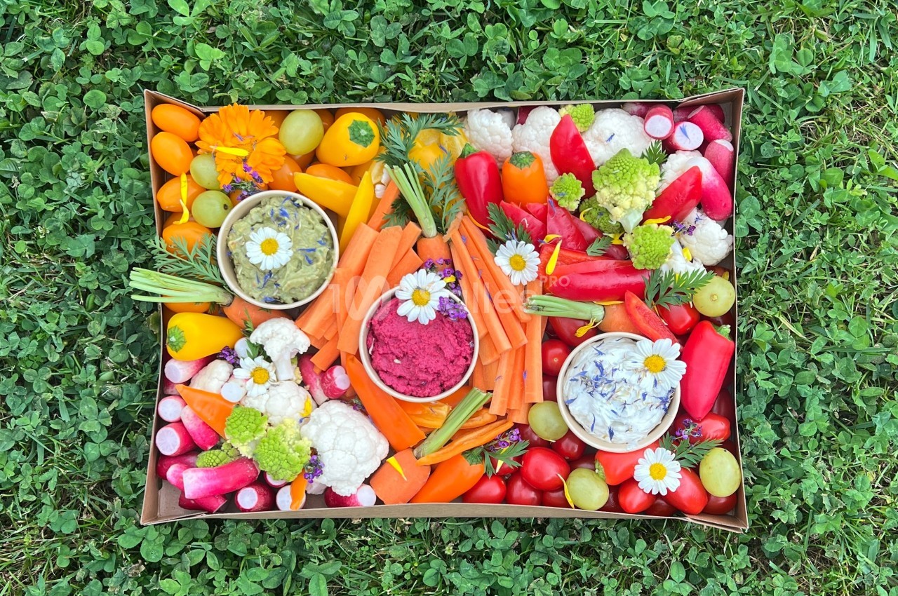 Plateau coloré de légumes frais (carottes, radis, poivrons) avec trois bols de dips, posé sur de l'herbe verte.