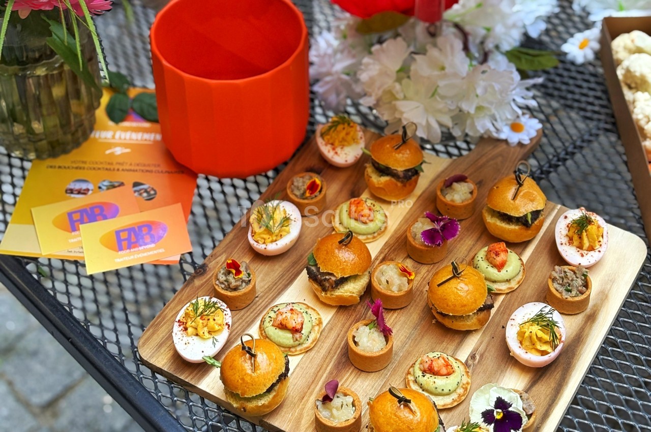 Plateau en bois avec des amuse-bouches variés, incluant mini-burgers, tartelettes et œufs garnis, posé sur une table en…