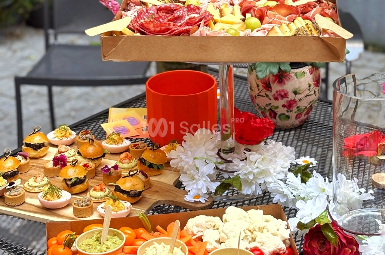 Plateau de charcuterie, mini-burgers et légumes frais disposés sur une table décorée de fleurs.