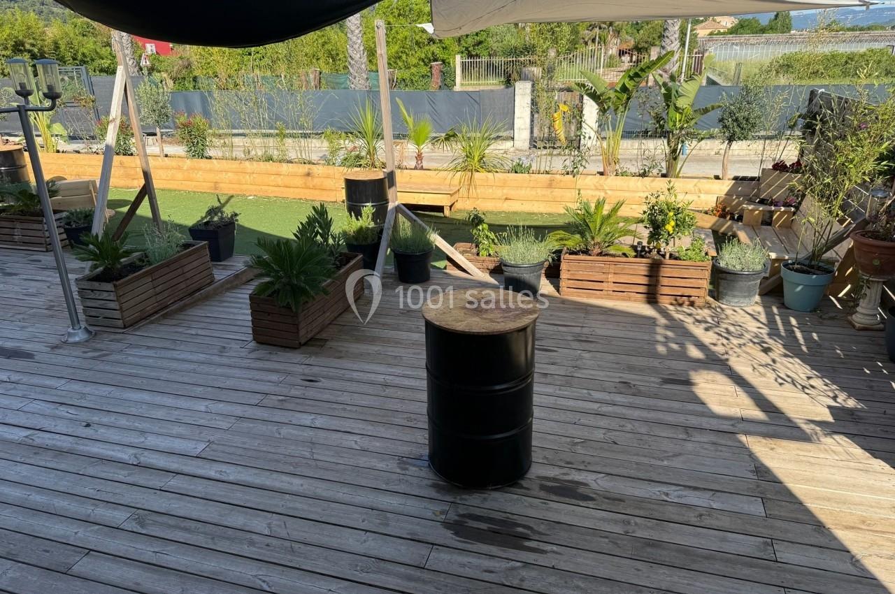 Terrasse en bois avec des plantes en pots, un tonneau central et des voiles d'ombrage, près d'un espace extérieur verdoyant.
