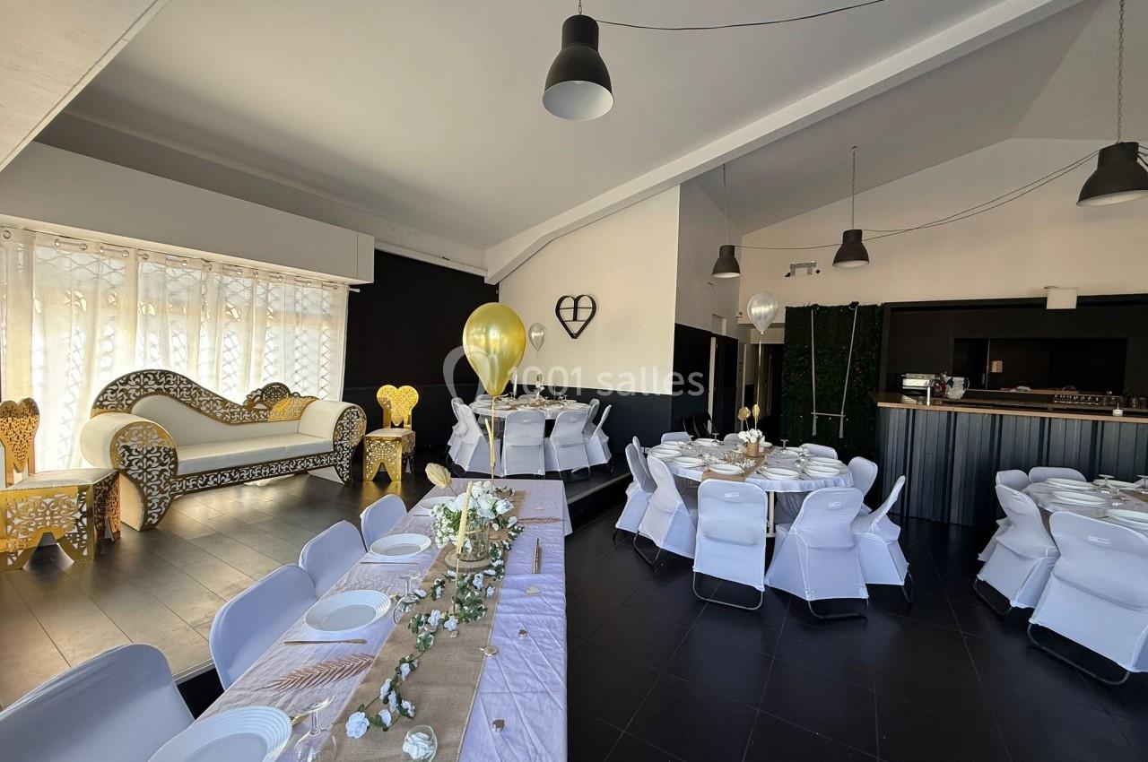 Salle de réception décorée avec des tables dressées, un canapé doré, des ballons et un espace bar en arrière-plan.