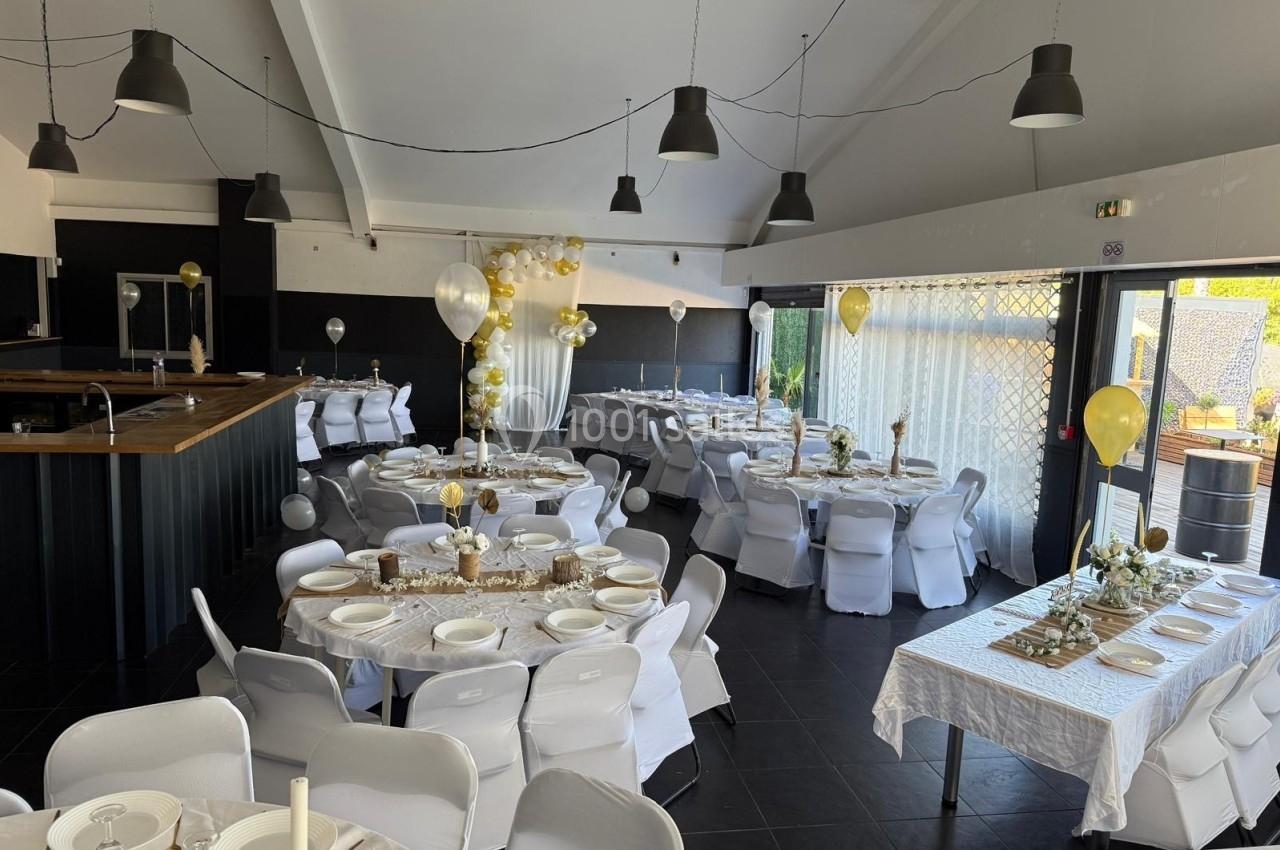 Salle de réception décorée pour un événement, avec tables dressées, nappes blanches, ballons et guirlandes dorées.