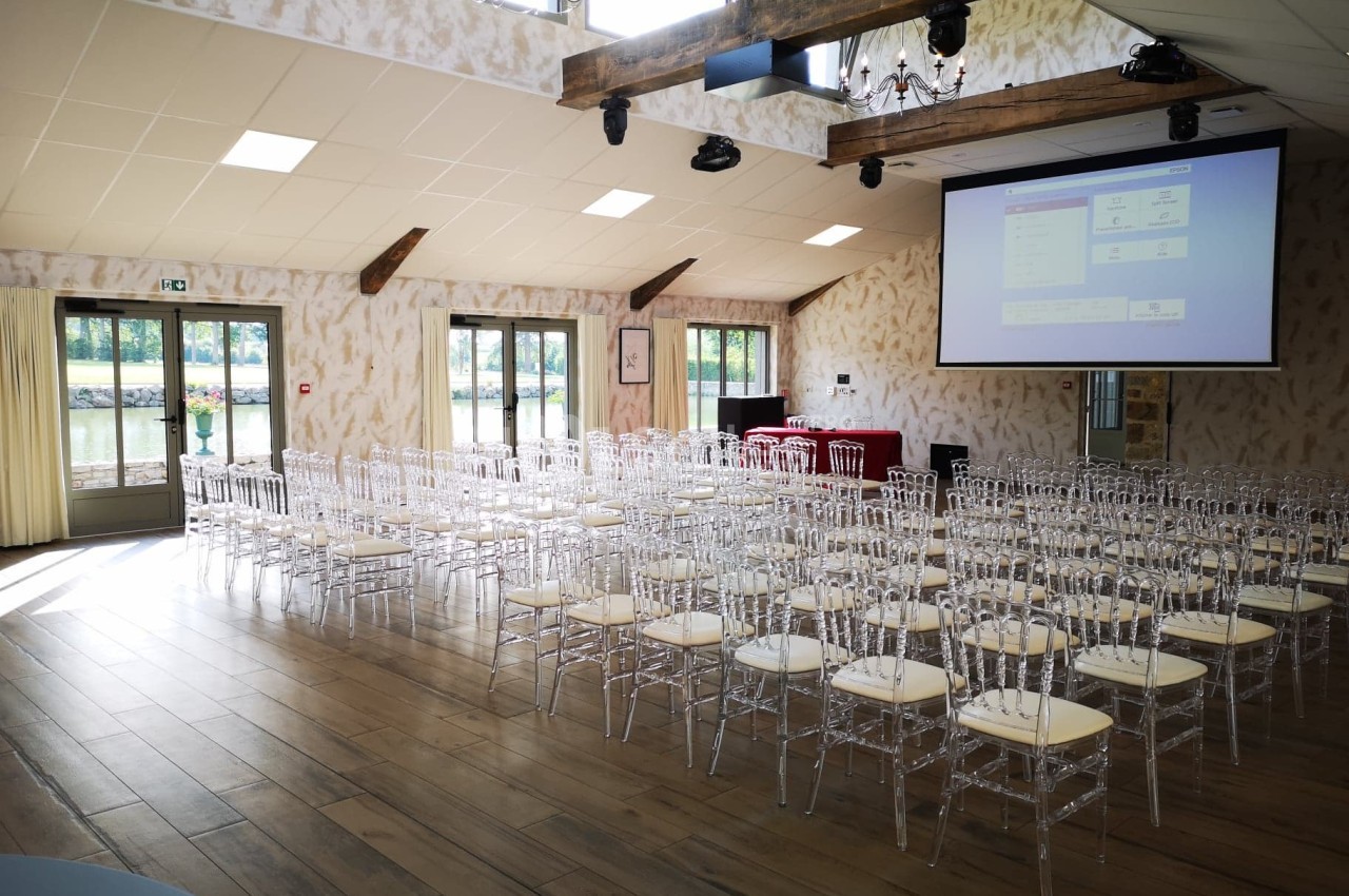Salle lumineuse aménagée avec des chaises transparentes, un écran de projection et des fenêtres donnant sur l'extérieur.