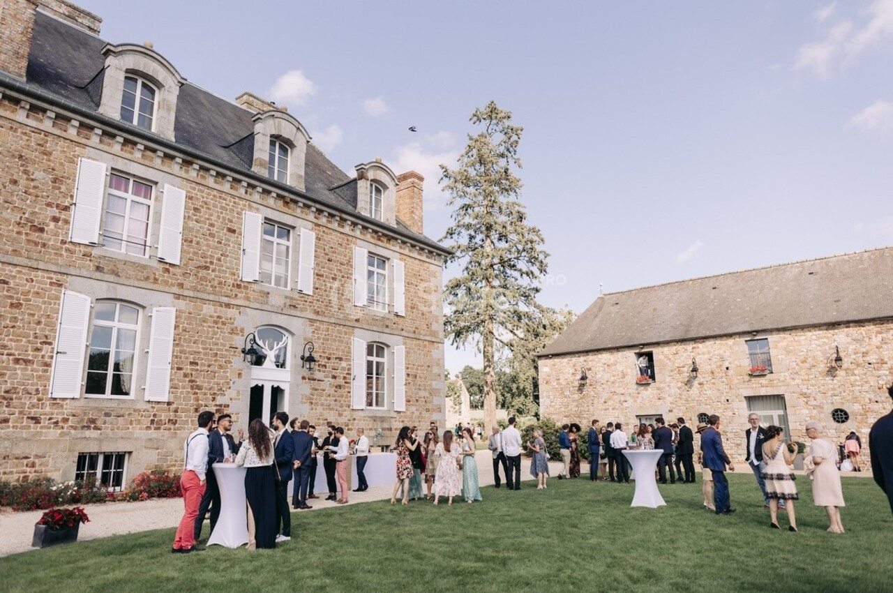Des invités discutent debout dans un jardin devant une grande maison en pierre et une grange sous un ciel dégagé.