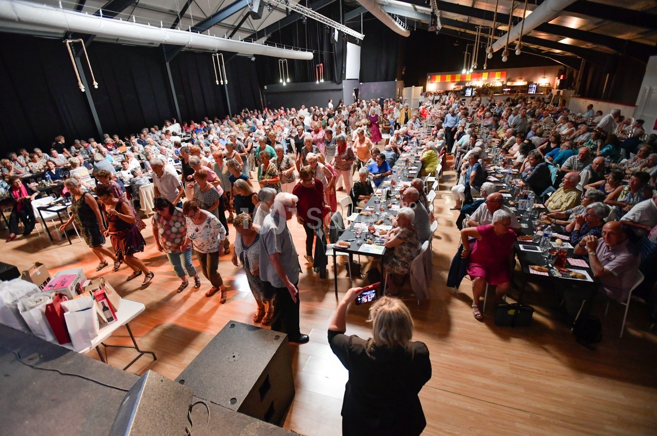 Salle comble avec des personnes assises et debout lors d'un événement convivial dans un espace intérieur lumineux.