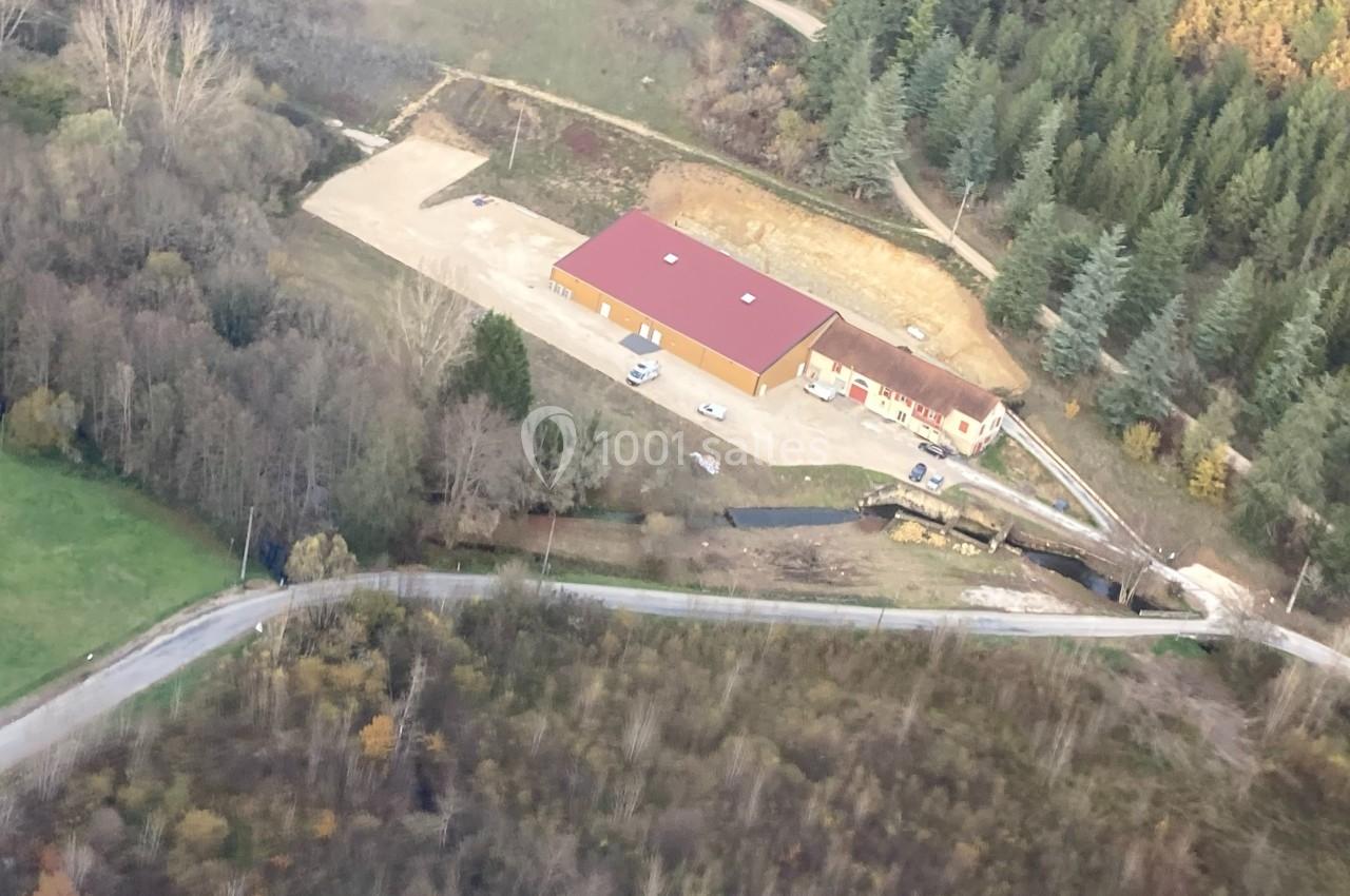 Vue aérienne d'un bâtiment avec un toit rouge entouré de végétation, d'une route et d'un petit cours d'eau.