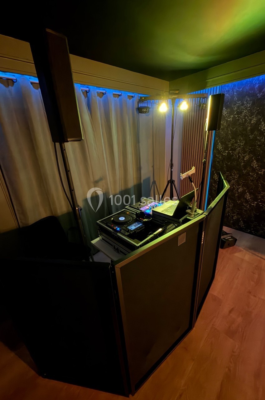 Poste de DJ avec platines, table de mixage et éclairage, installé dans une pièce sombre avec rideaux et murs texturés.