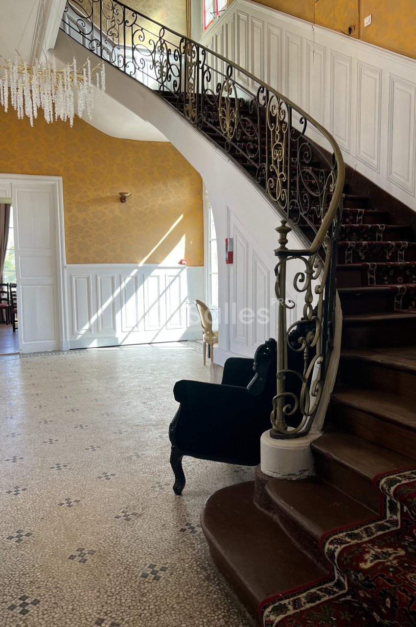 Escalier en bois avec rampe en fer forgé dans un hall lumineux, sol en mosaïque et lustre suspendu.