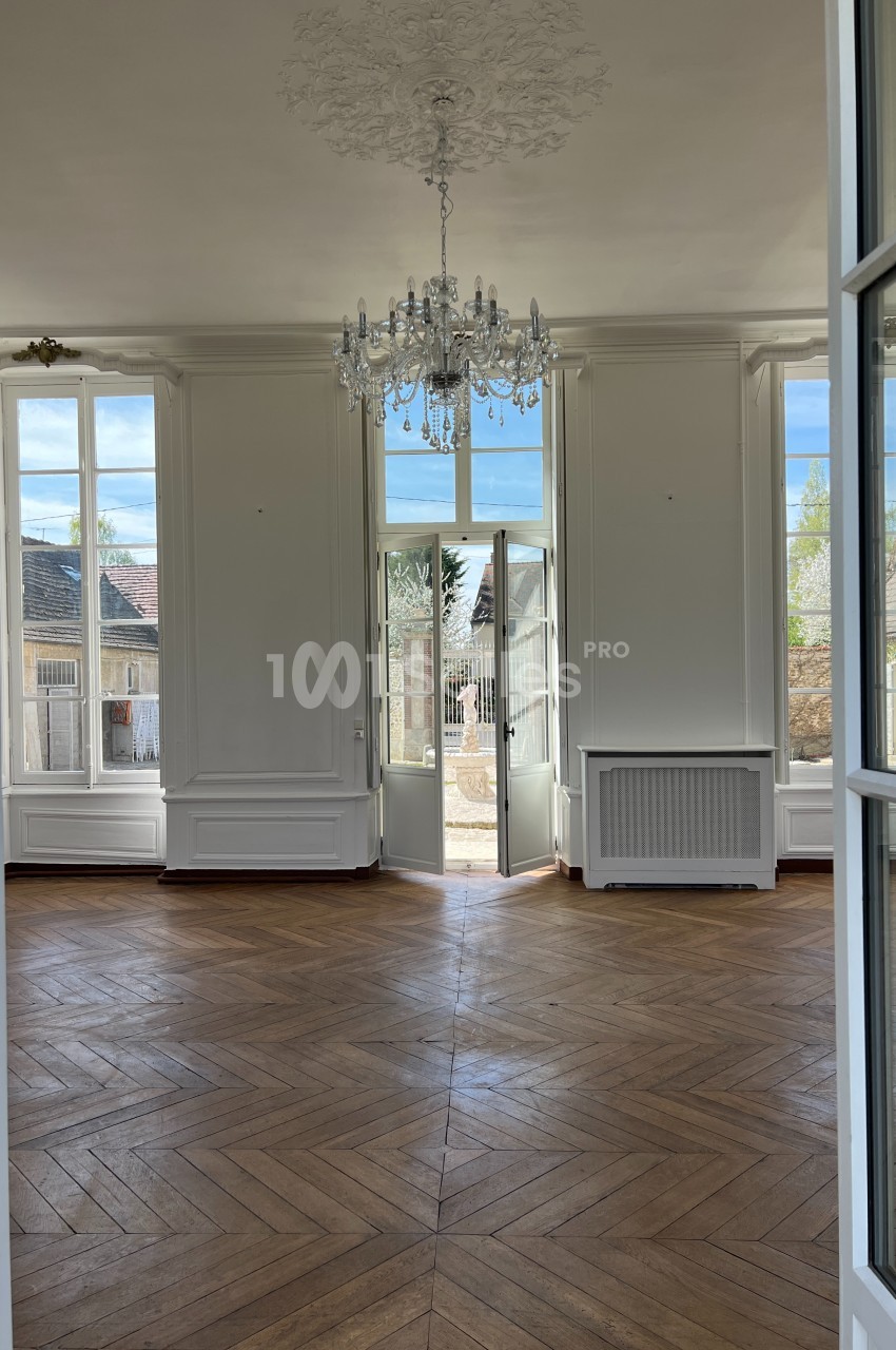 Salle lumineuse avec parquet en bois, grandes fenêtres, porte-fenêtre ouverte sur un jardin et lustre au plafond.