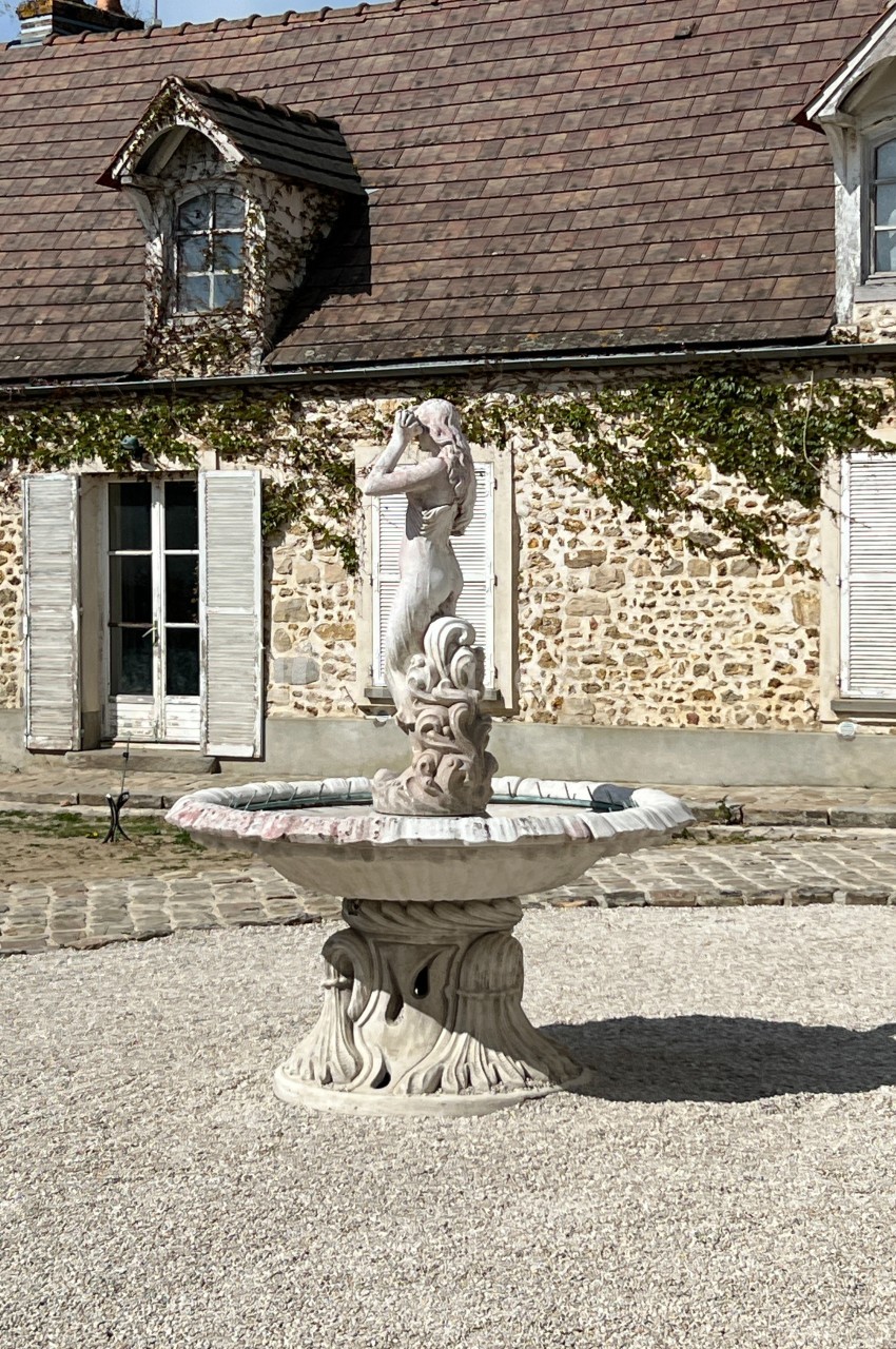 Fontaine en pierre ornée d'une statue, devant une maison en pierre avec volets blancs et toit en tuiles.