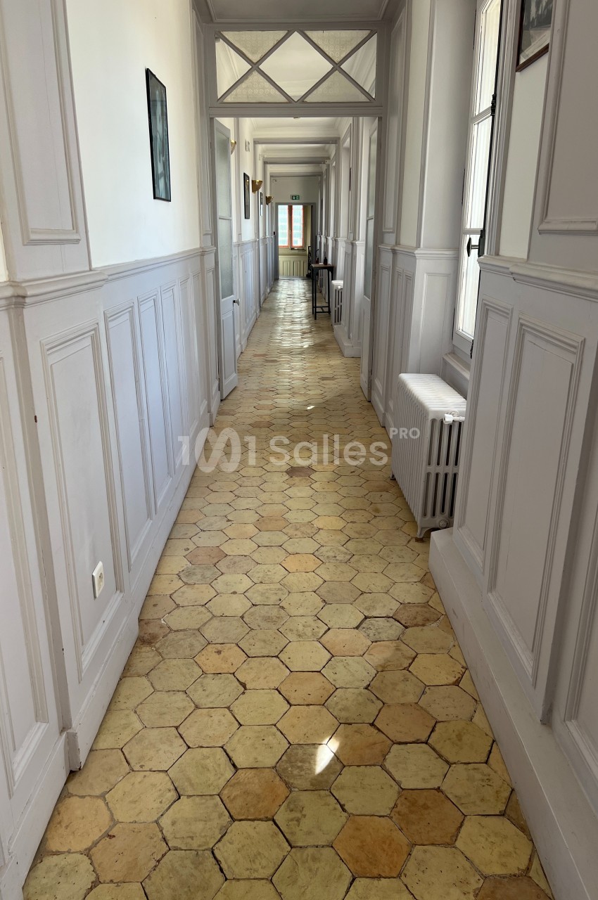 Couloir lumineux avec sol en carreaux hexagonaux, murs blancs et fenêtres laissant entrer la lumière naturelle.