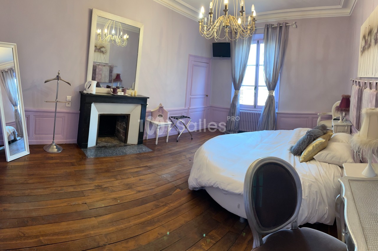 Chambre lumineuse avec parquet, lit double, cheminée, miroir, bureau et lustre, décorée dans des tons pastel.