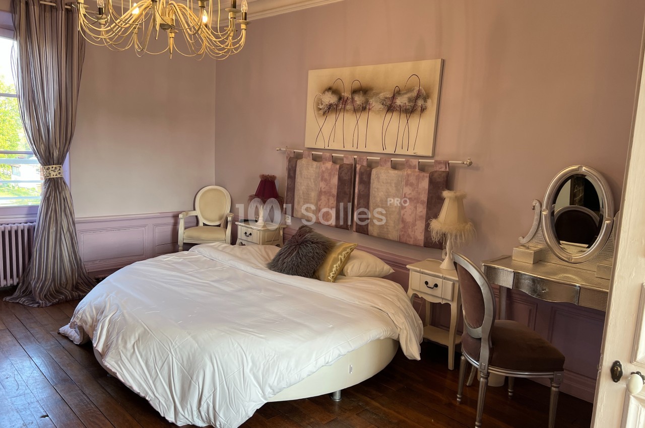 Chambre élégante avec lit rond, mobilier classique, murs mauves et lustre doré, éclairée par une fenêtre.