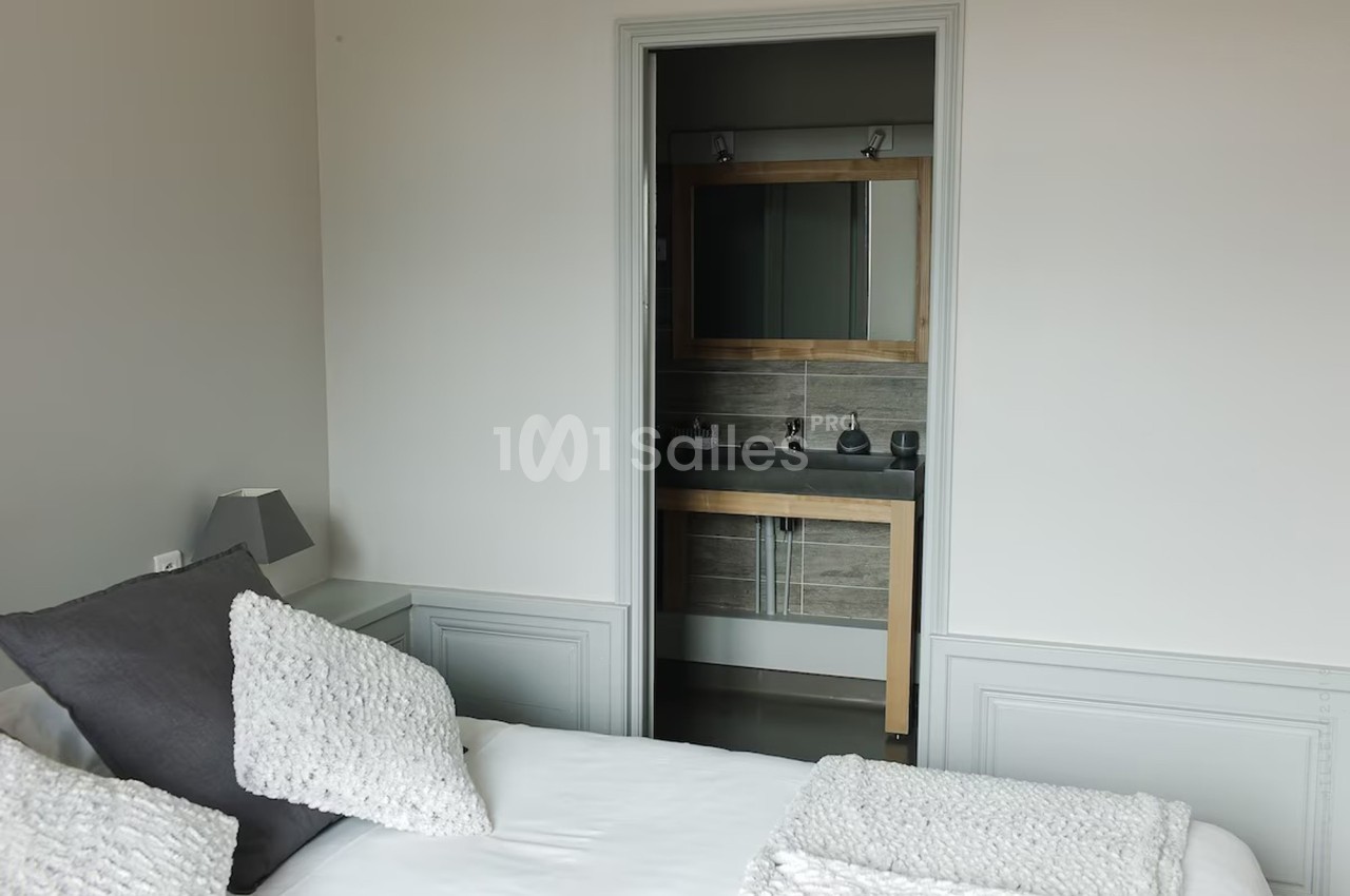 Vue d'une chambre avec un lit blanc et des coussins gris, donnant sur une salle de bain avec un miroir et un lavabo.