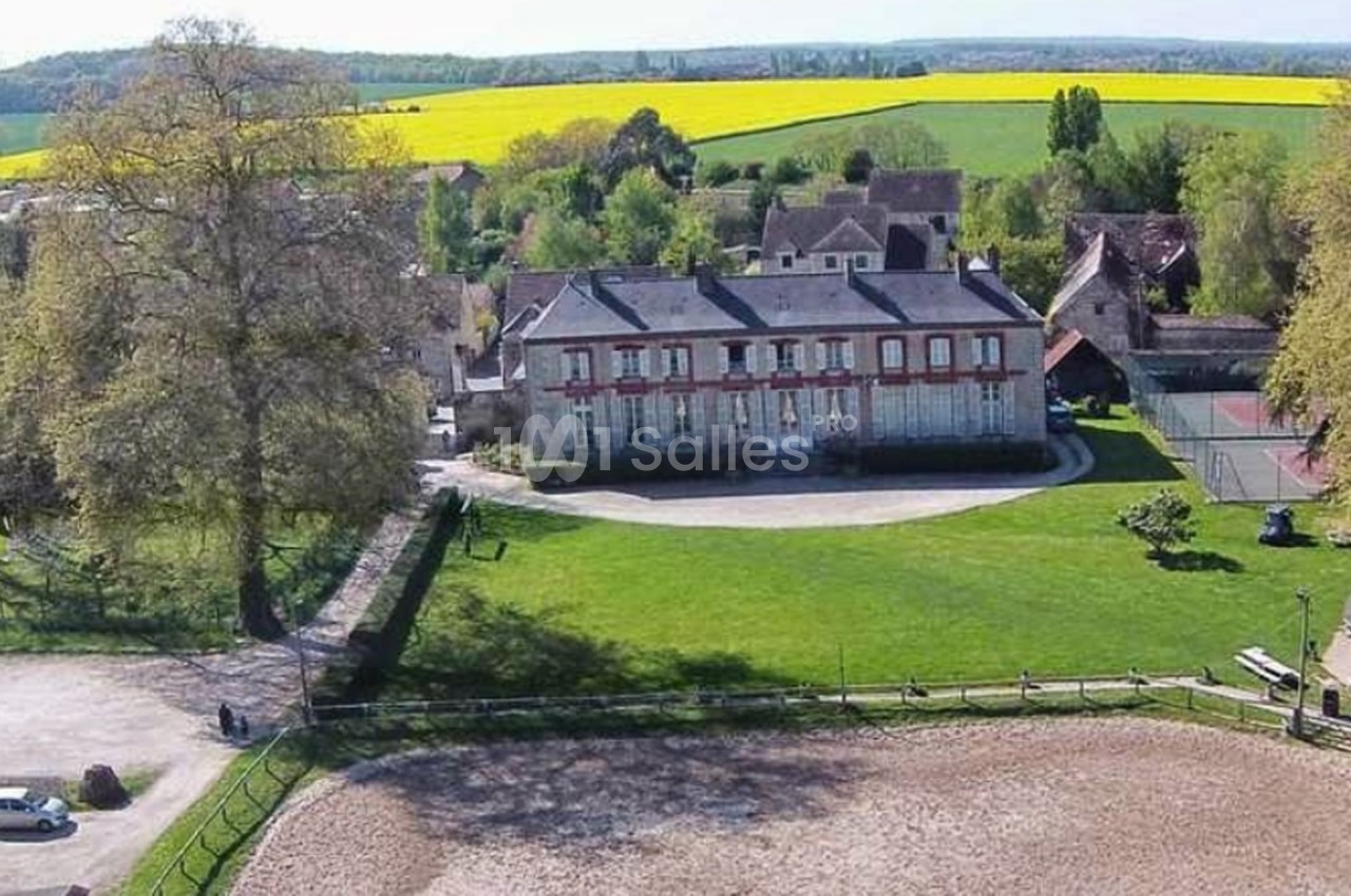 Vue aérienne d'un domaine avec un bâtiment principal, une cour, des arbres et des champs de colza en arrière-plan.