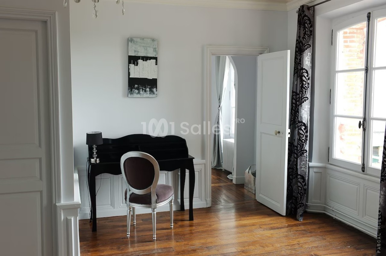 Bureau avec un petit secrétaire noir, chaise argentée, lampe, et fenêtre laissant entrer la lumière naturelle.
