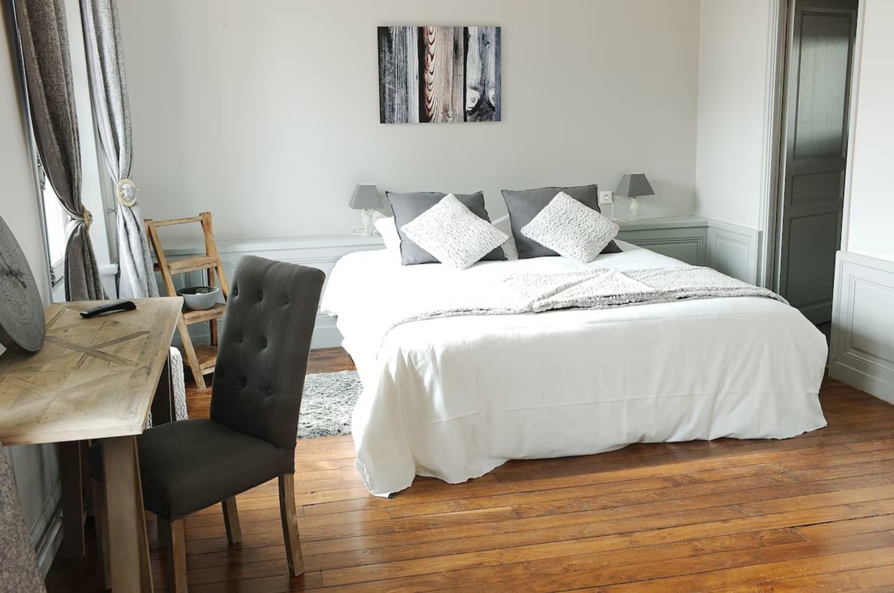 Chambre lumineuse avec lit double, draps blancs, coussins gris, bureau en bois et parquet en bois clair.