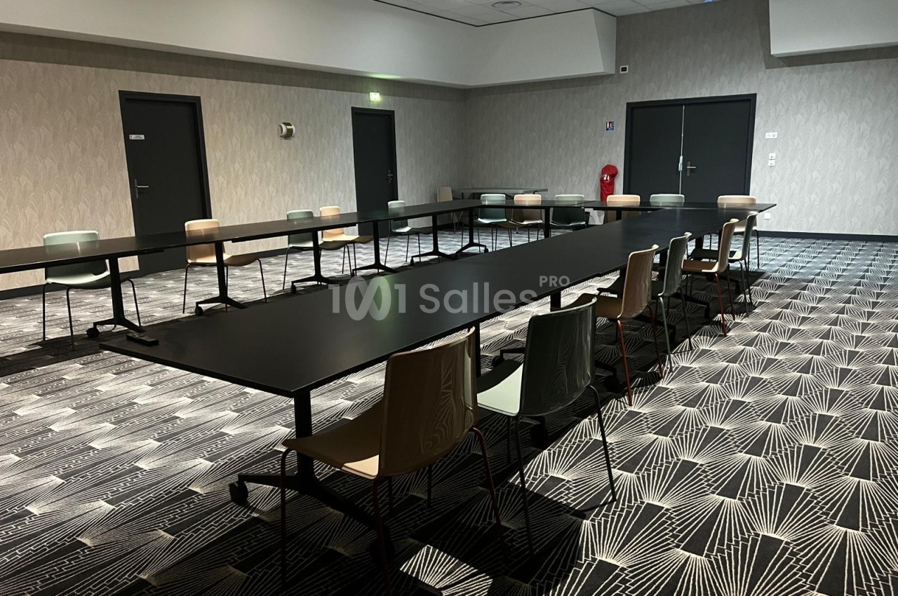 Salle de réunion vide avec tables noires disposées en U, chaises colorées et sol à motifs géométriques.