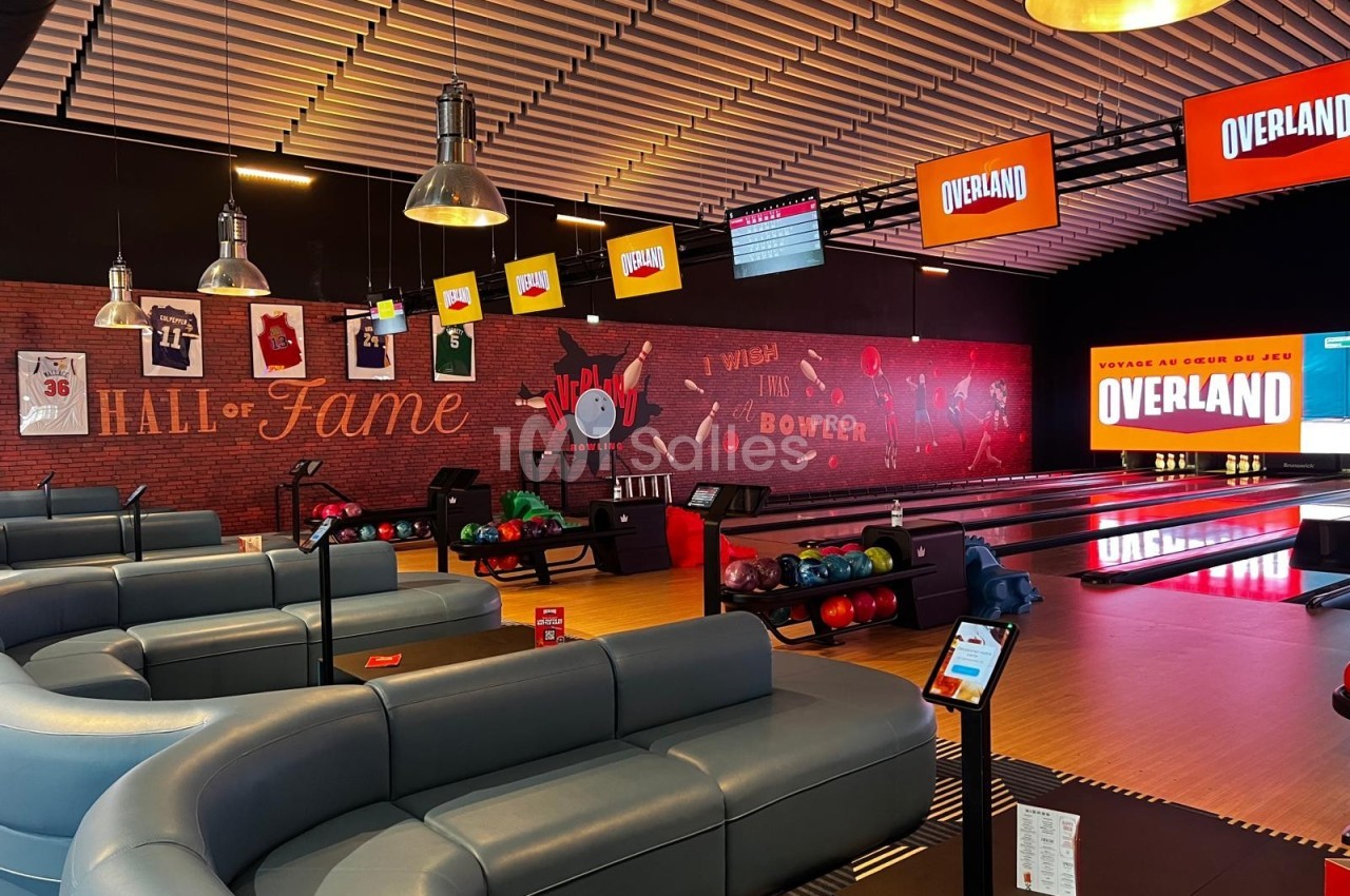 Salle de bowling moderne avec pistes éclairées, canapés gris, écrans suspendus et décorations murales sportives.