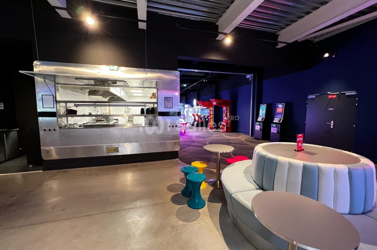 Espace intérieur avec coin salon moderne, tables colorées, cuisine ouverte et machines d'arcade en arrière-plan.