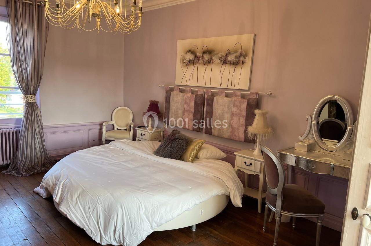 Chambre élégante avec murs mauves, lit rond, parquet en bois, lustre doré et coiffeuse avec miroir.