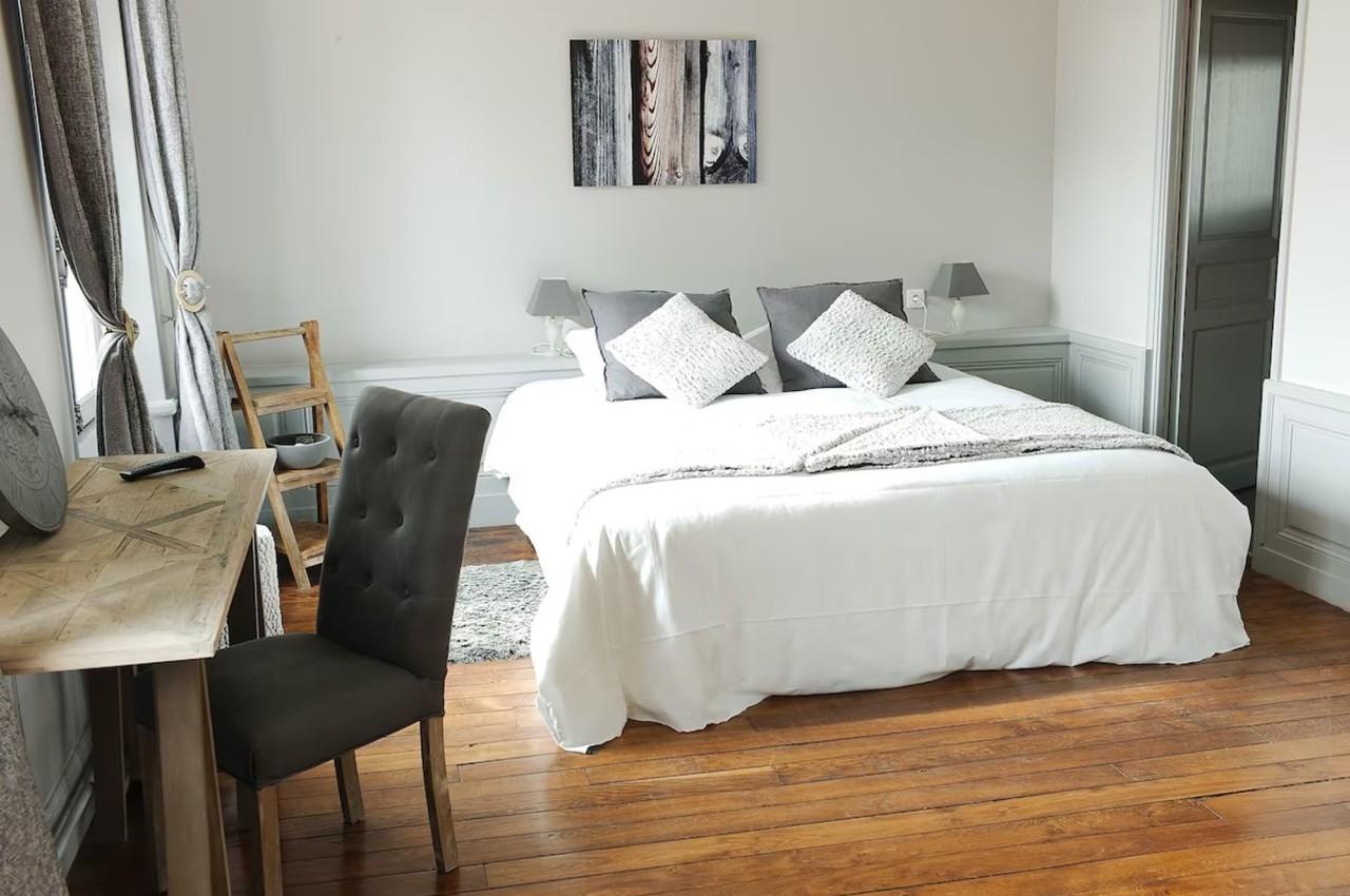 Chambre lumineuse avec lit double, draps blancs, coussins gris, bureau en bois et parquet en bois clair.