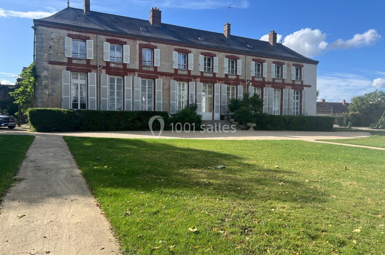 Façade d'un grand bâtiment en pierre avec volets blancs, entouré de pelouses et d'un ciel dégagé.