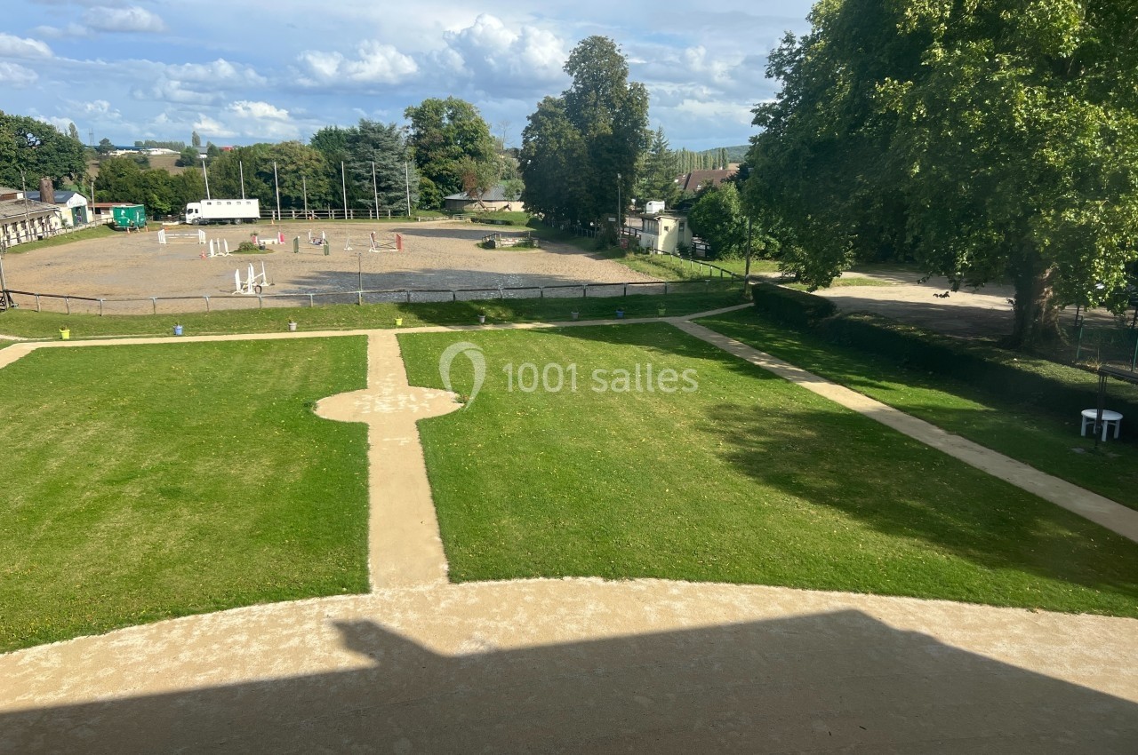 Vue sur un grand jardin avec pelouses, allées en gravier et un terrain d'équitation en arrière-plan sous un ciel…