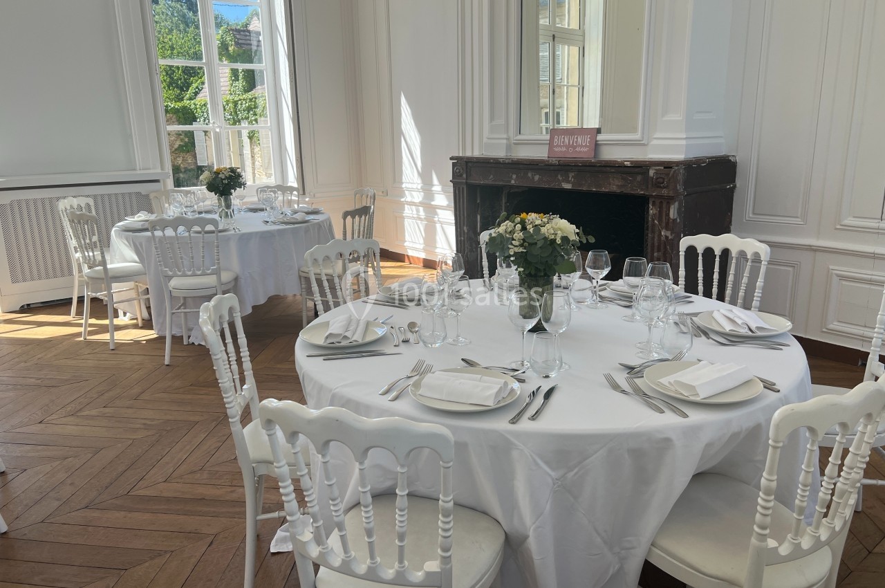 Salle lumineuse avec tables rondes dressées pour un repas, décorées de fleurs, dans un intérieur élégant avec cheminée.