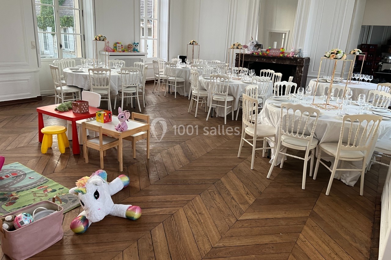 Salle de réception lumineuse avec tables dressées et espace de jeux pour enfants au premier plan.