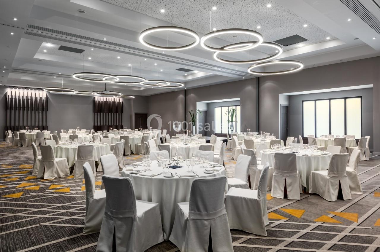 Salle de réception moderne avec tables rondes dressées, chaises blanches et éclairage circulaire suspendu.