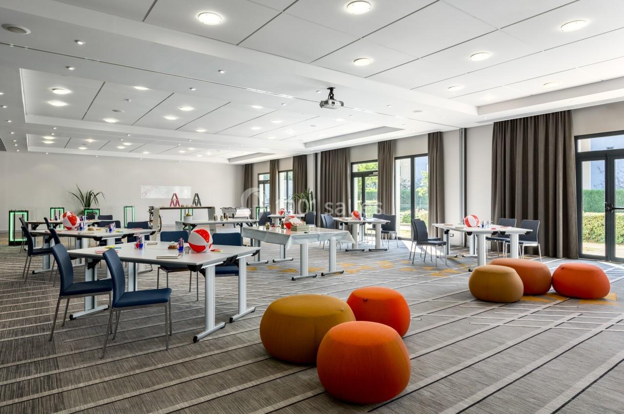 Salle de réunion lumineuse avec tables, chaises, poufs colorés et grandes fenêtres donnant sur un espace vert.