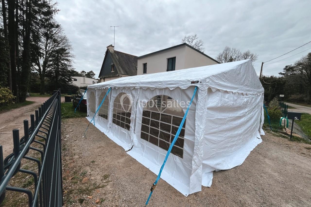 Tente blanche installée dans une cour gravillonnée près d'une maison, avec des arbres en arrière-plan.