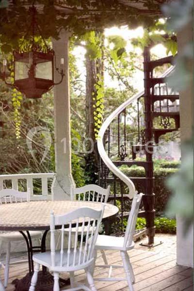 Escalier en colimaçon en métal noir près d'une terrasse en bois avec table et chaises blanches, entourée de verdure.