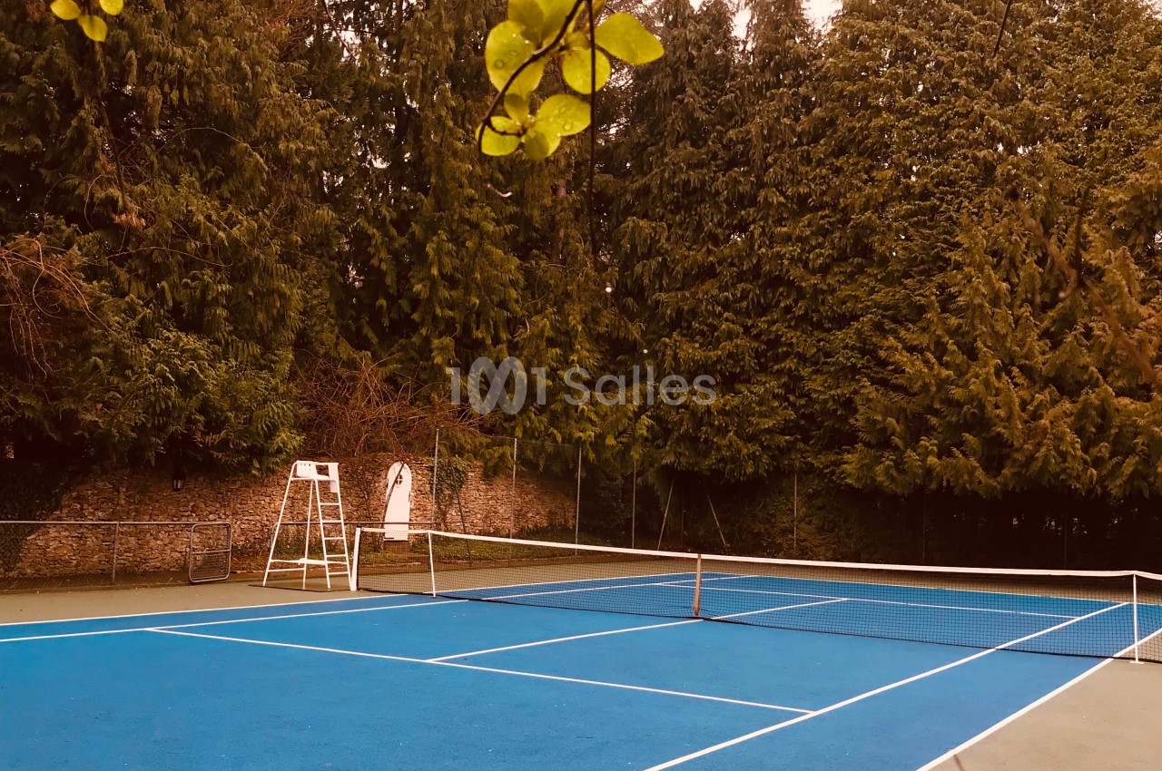Terrain de tennis extérieur avec revêtement bleu, entouré d'arbres et équipé d'une chaise d'arbitre blanche.